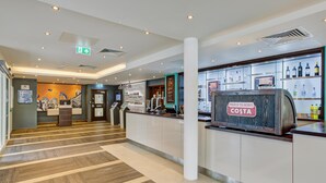 Um hall de hotel com uma mesa de recepção, um balcão com o aviso 'Proud to Serve Costa', uma exposição de bebidas engarrafadas e um mural na parede.