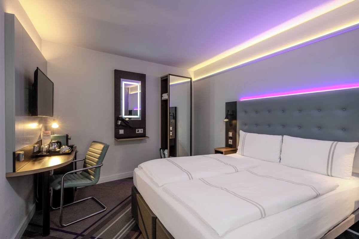 A hotel room with a bed, a desk, a chair, a mirror, and a wall-mounted TV.