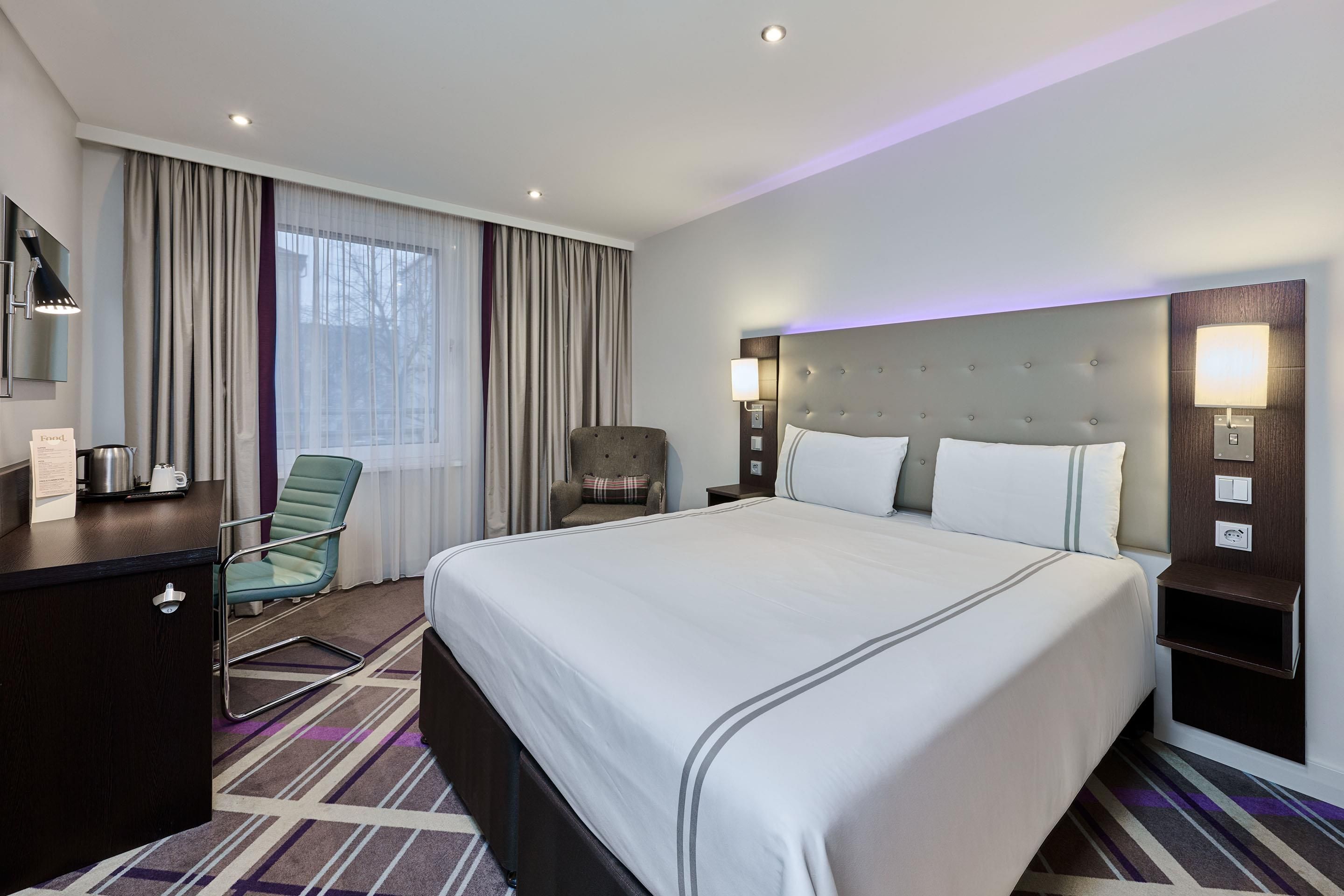 A hotel room with a large bed, a desk, a chair, a nightstand, and a window with curtains.