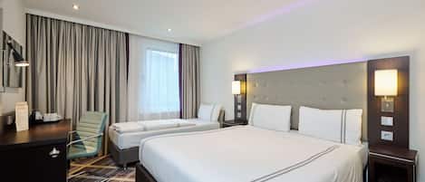 A hotel room with a large bed, a smaller bed, a desk with a chair, a bedside table, and a window with curtains.