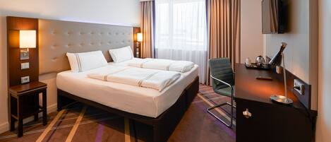 Une chambre d’hôtel avec un grand lit, un bureau avec une lampe, une chaise, une télévision et une fenêtre avec des rideaux.