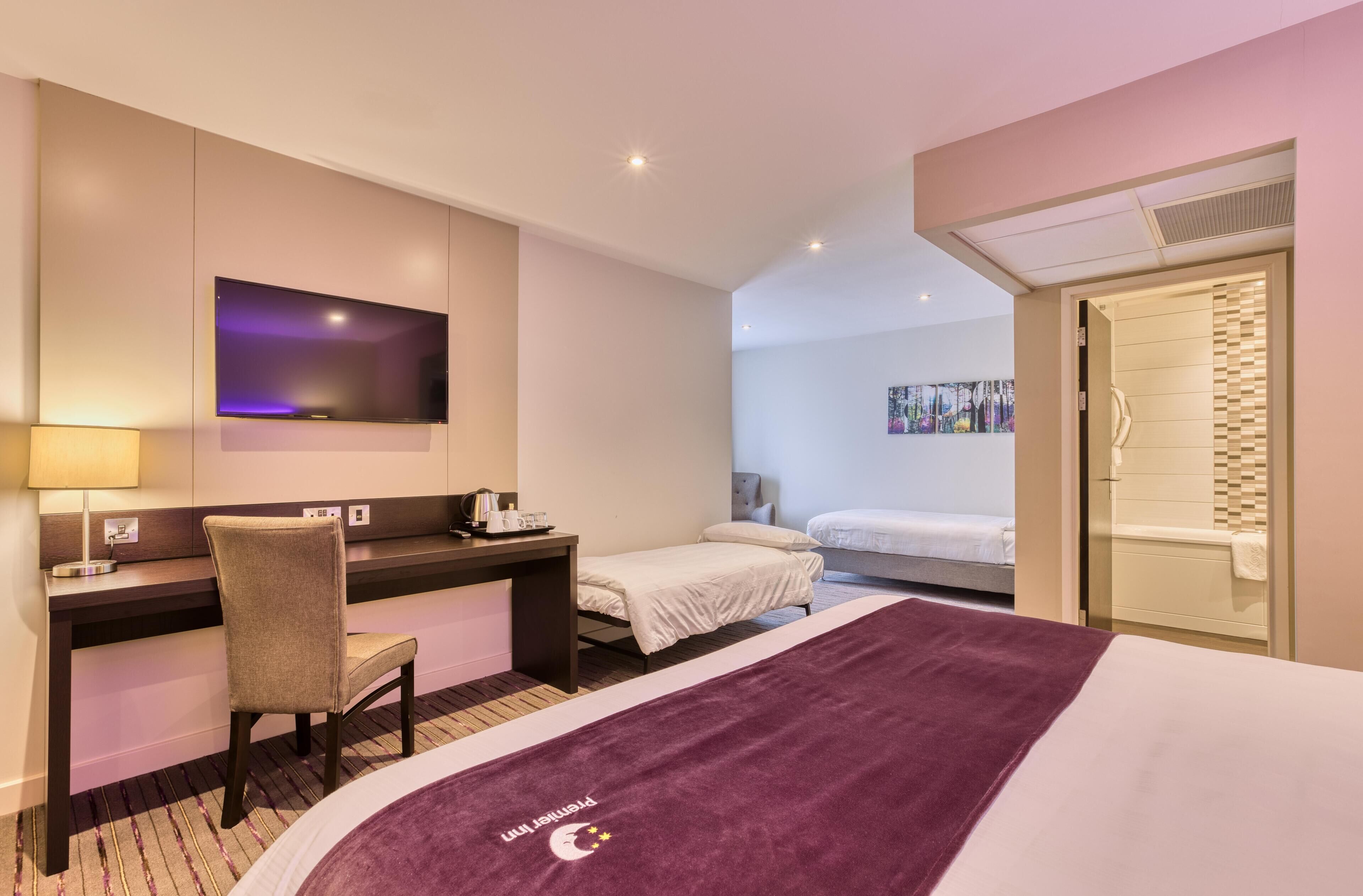 A hotel room with a bed, a desk with a chair, a TV, a shower, and a painting on the wall.