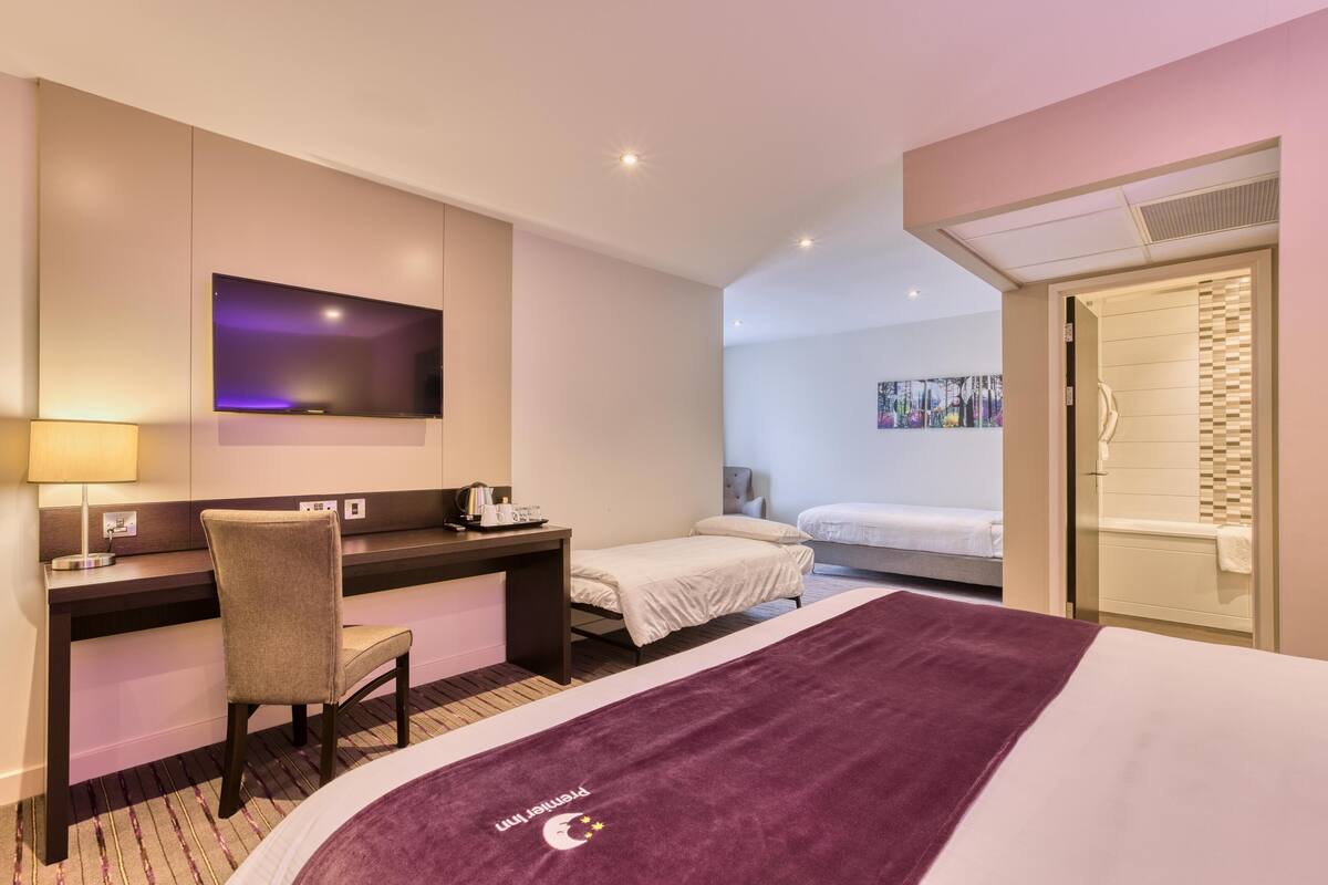 A hotel room with a bed, a desk with a chair, a TV, a shower, and a painting on the wall.