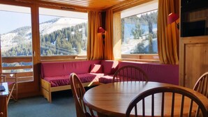 Una cabaña acogedora con interior de madera, una mesa redonda de madera y un banco tapizado rojo con vistas a montañas nevadas.