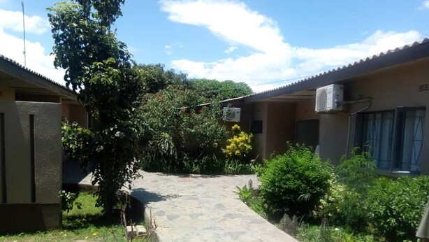 A courtyard with a paved walkway, surrounded by greenery and two buildings with air conditioning units. - Pakali Lodges (Livingstone)