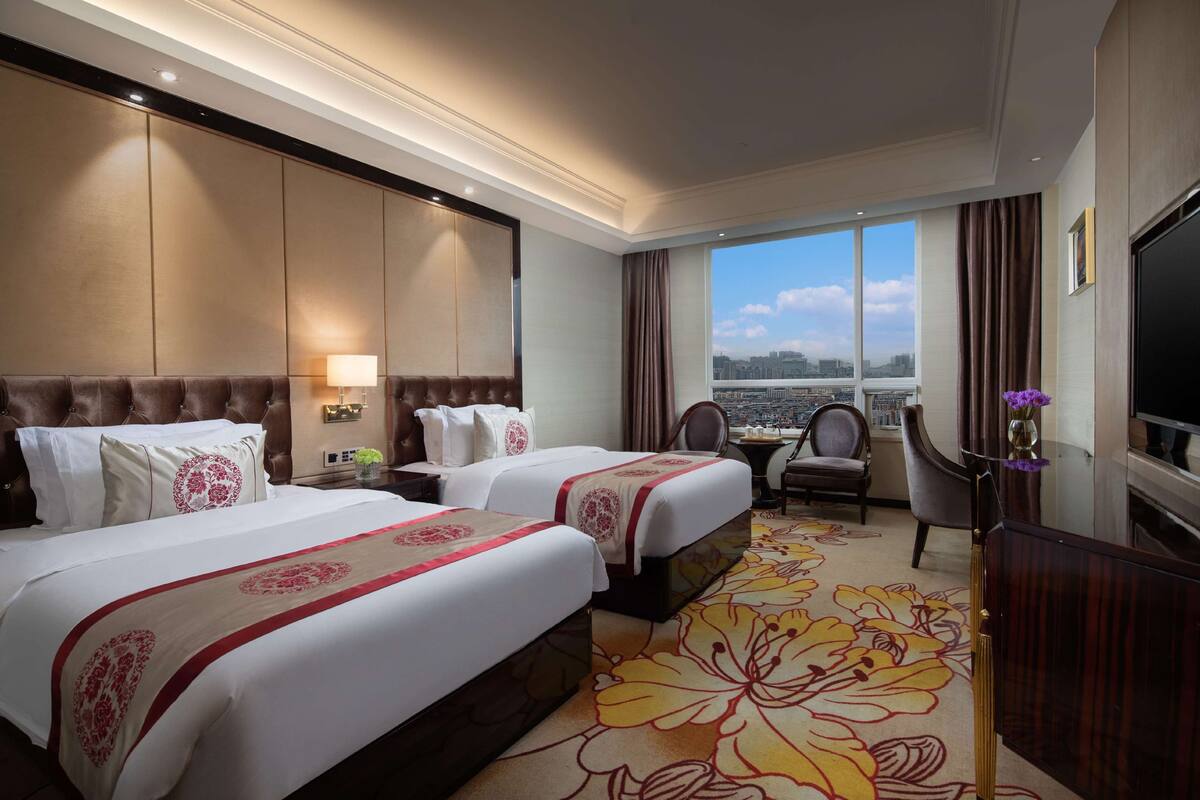 Ein Hotelzimmer mit zwei Betten, einem großen Fenster mit Blick auf die Stadt, einem Fernseher und einem Teppich mit Blumenmuster.