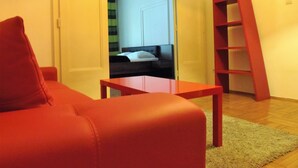 Una habitación con un sofá rojo, una mesita de madera, una escalera roja y una cama con una cortina a rayas al fondo.