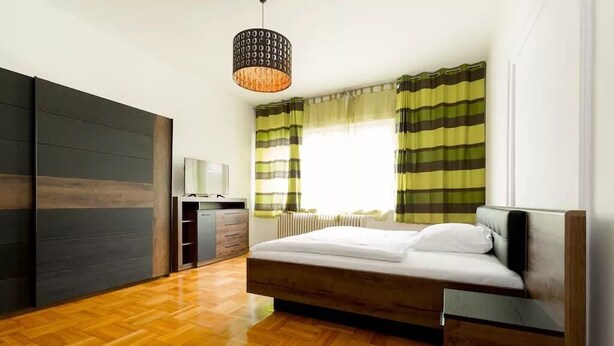 Un dormitorio moderno con suelo de madera, una cama con ropa de cama blanca, una mesita de noche, un armario y una ventana con cortinas a rayas.