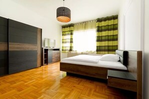 A modern bedroom with a wooden floor, a bed with white bedding, a bedside table, a wardrobe, and a window with striped curtains. - Budapest Promenade Green (Budapest)