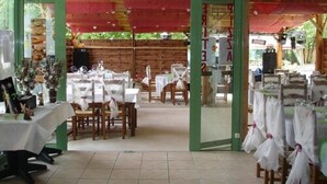 An indoor dining area with tables set for guests, a wooden wall in the background, and a glass door leading outside. - Camping la Garenne (Peyrignac)