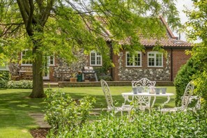 A stone and brick house with a garden patio featuring white metal chairs and a table. - Westgate Cottage (T) (4), Thornham, Norfolk (Thornham)