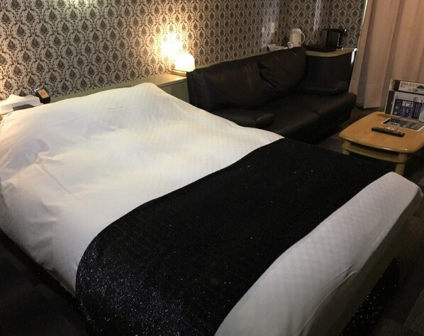 A hotel room with a bed, a black leather sofa, a small wooden table, and a nightstand with a lamp.