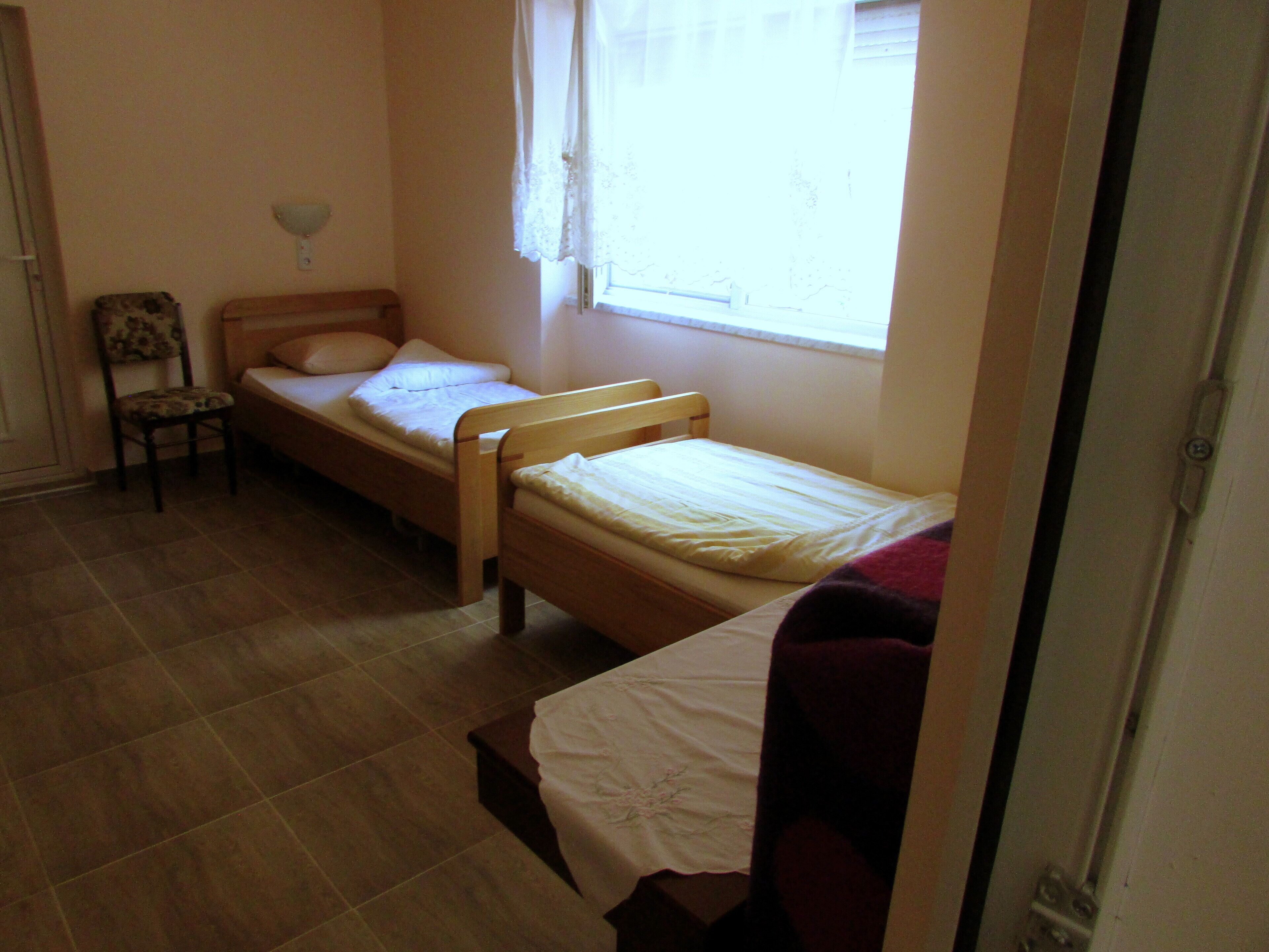 A small, single-room accommodation with two wooden beds, a chair, and a window with curtains.