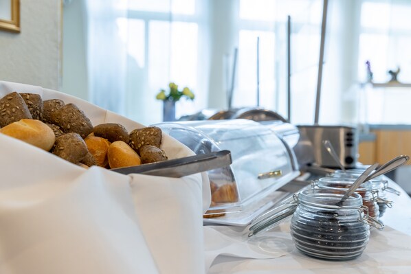 A breakfast buffet with various breads and pastries, a coffee machine, and condiment jars.