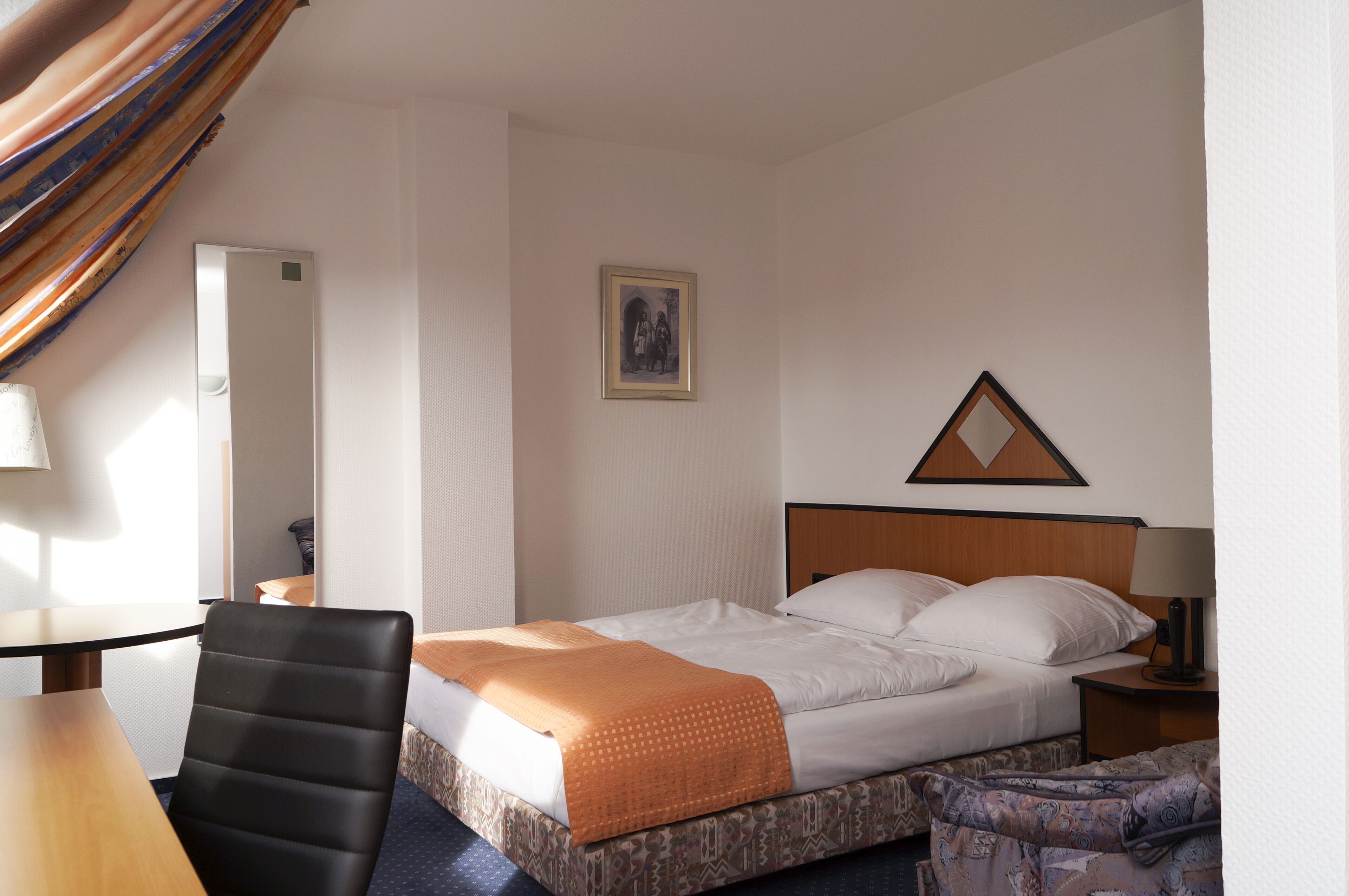 A hotel room with a bed, a chair, a table, a nightstand, and a picture on the wall.