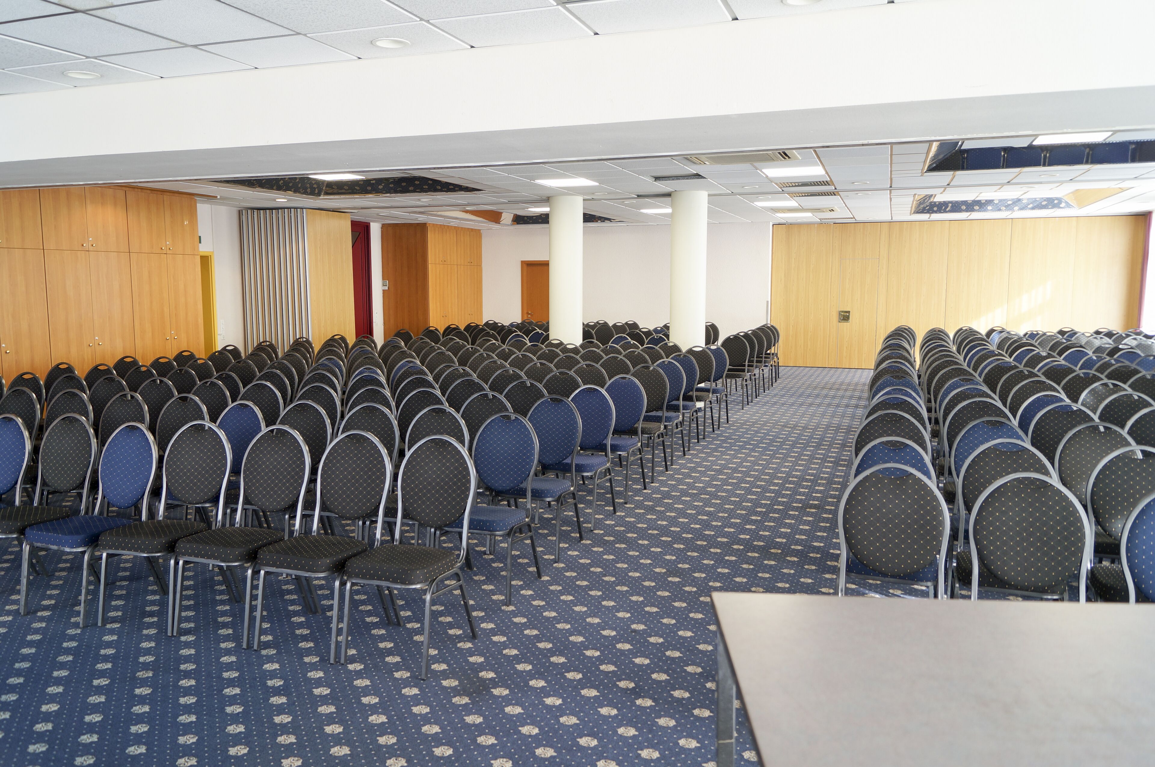 A conference room with rows of chairs and tables, wooden panel walls, and a patterned carpet.