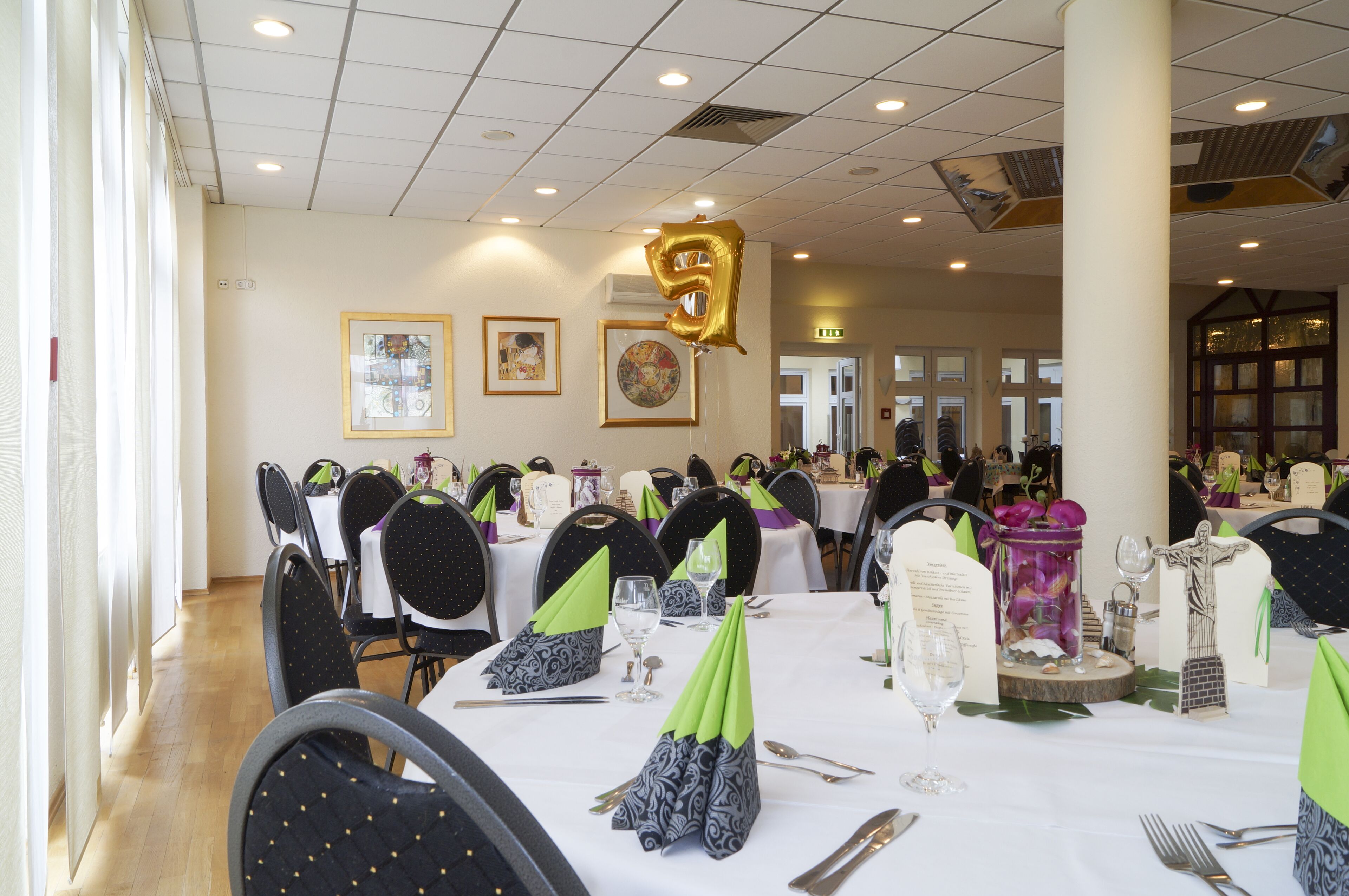 A banquet hall with tables set for a formal event, featuring green and black tablecloths, white napkins, and gold balloons.
