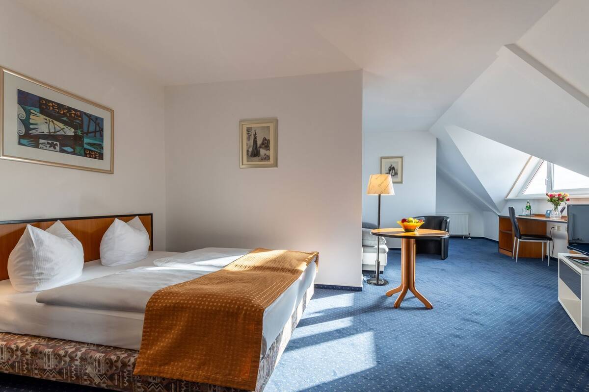A hotel room with a bed, a desk, a chair, a lamp, and a painting on the wall.
