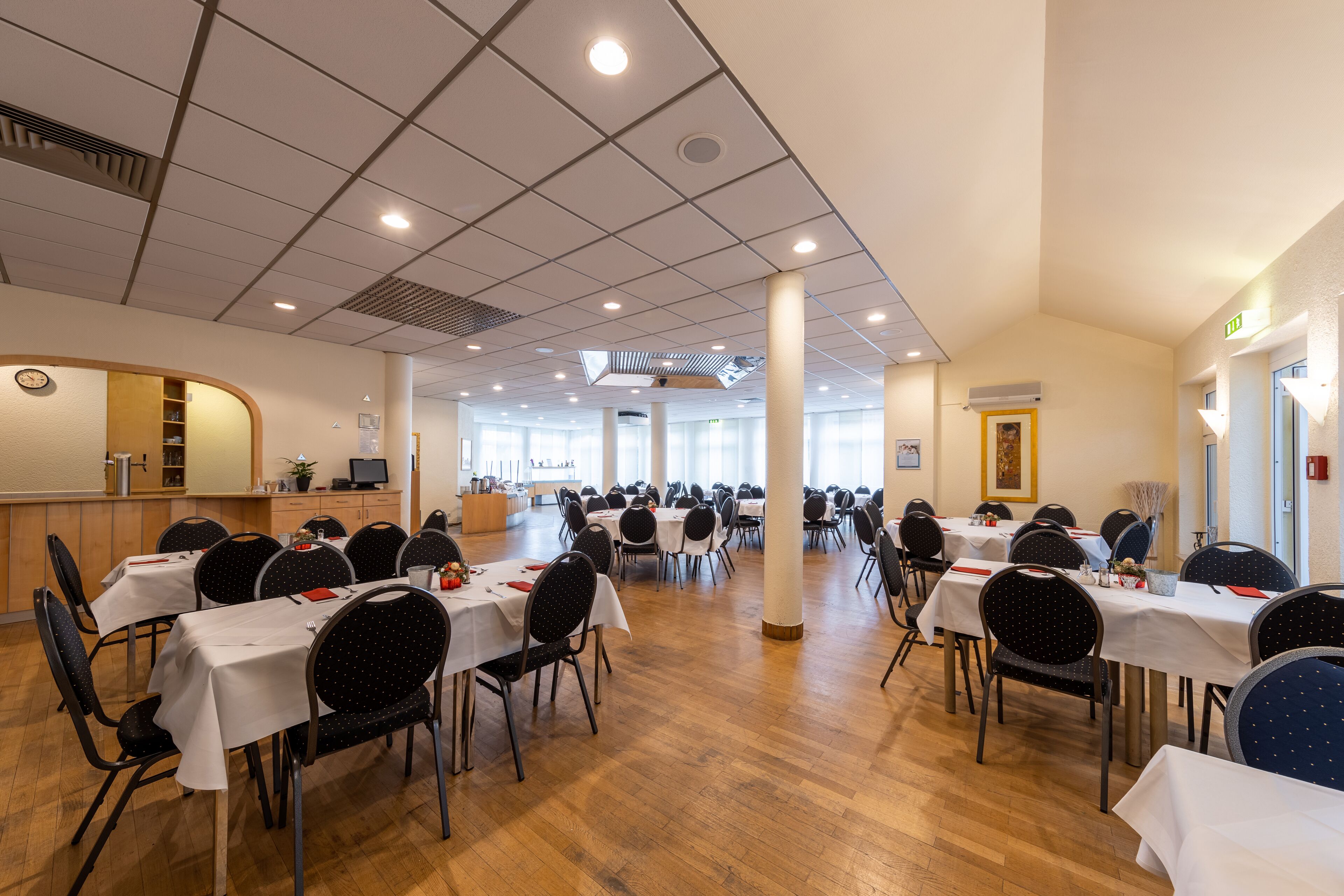 A spacious dining hall with multiple tables and chairs, a reception area, and large windows allowing natural light.