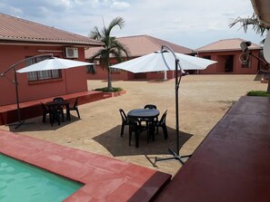 A pool area with a red-tiled deck, two outdoor tables with chairs, and white umbrellas providing shade. - Fallsview Apartments (Livingstone)