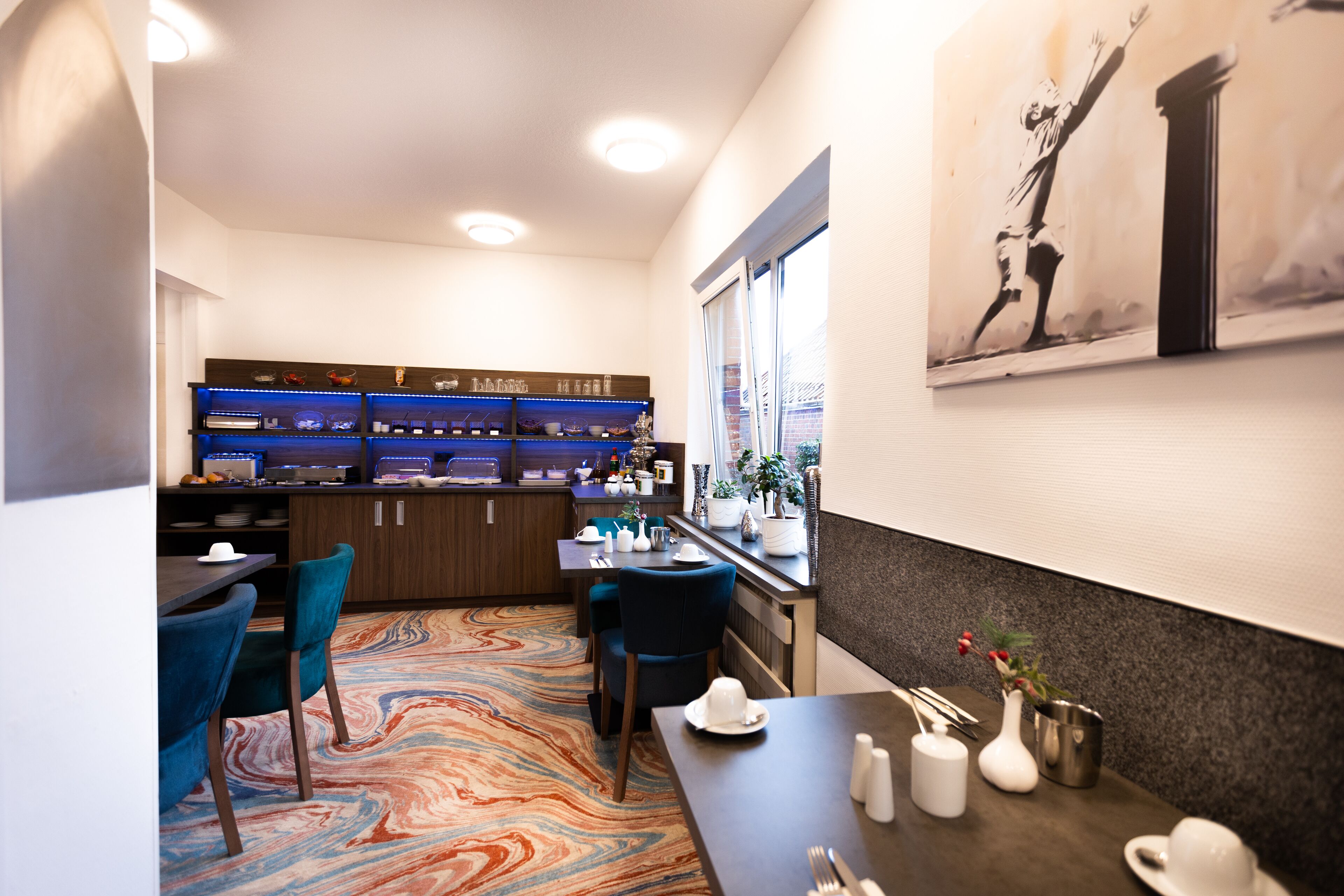A hotel room with a dining area, a kitchen with blue lighting, a colorful carpet, and a painting on the wall.