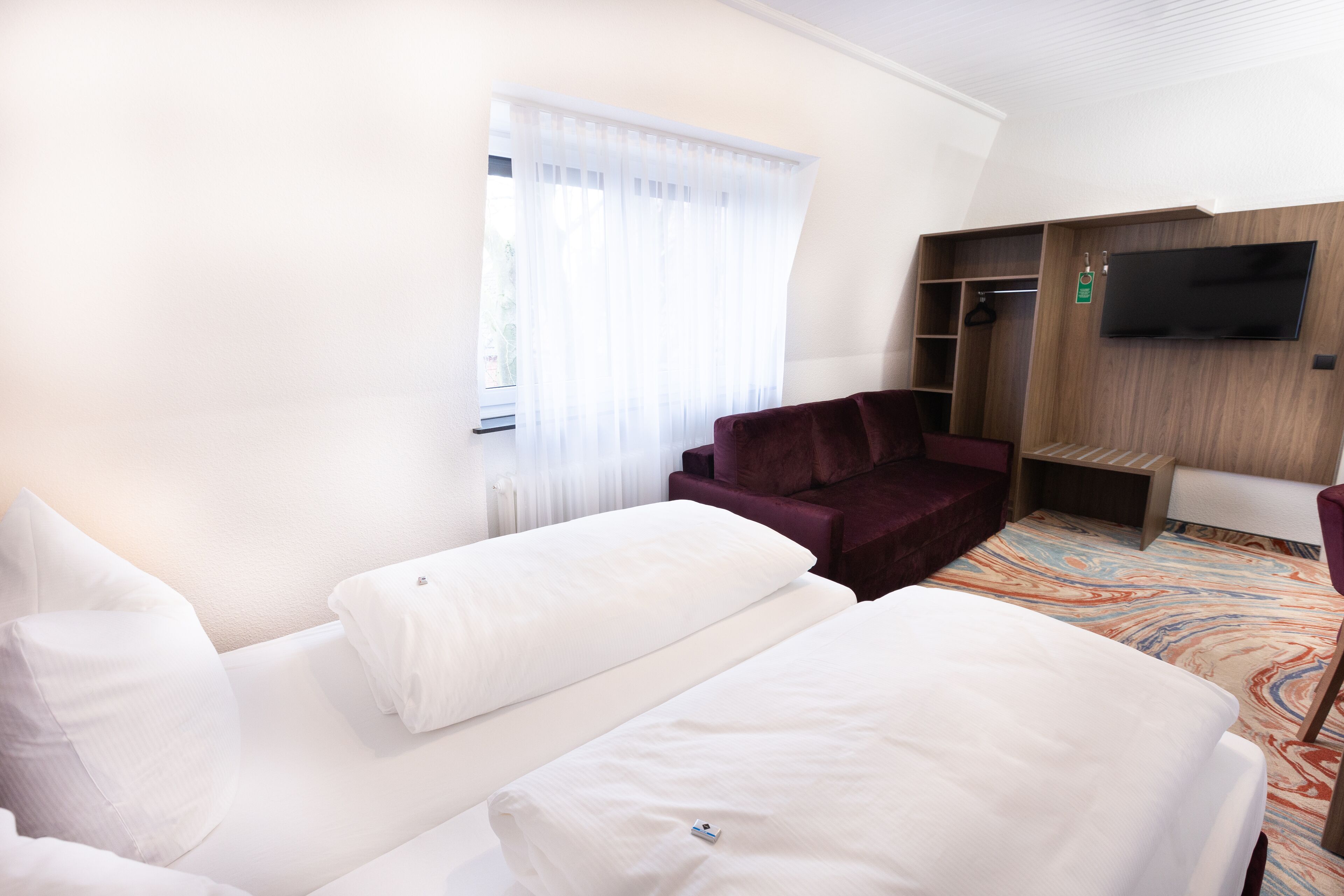 Habitación de hotel con dos camas, una zona de estar con sofá y sillones, un televisor montado en la pared y una ventana con cortinas traslúcidas.