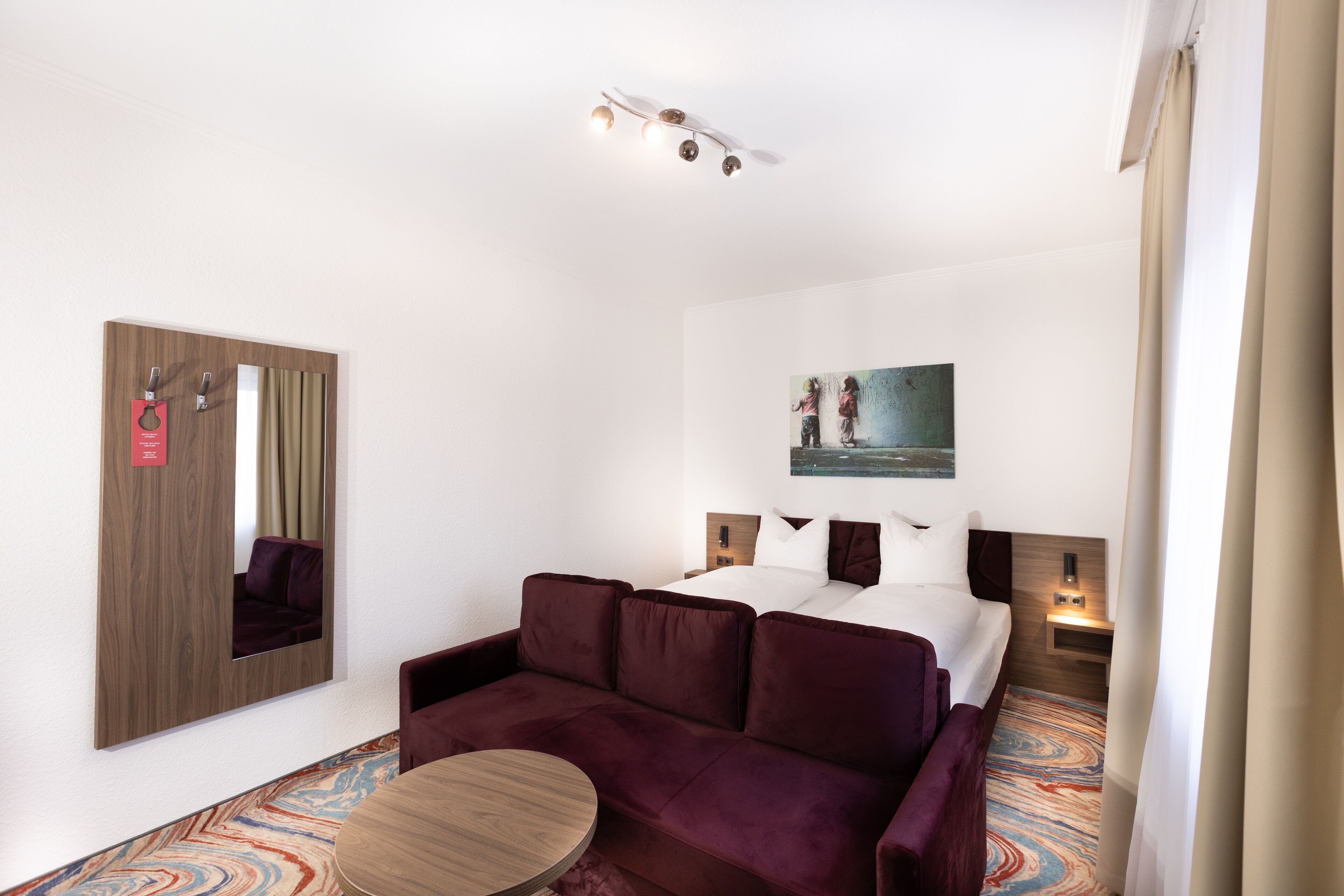 A hotel room with a bed, a sofa, a round table, a mirror, and a painting on the wall.