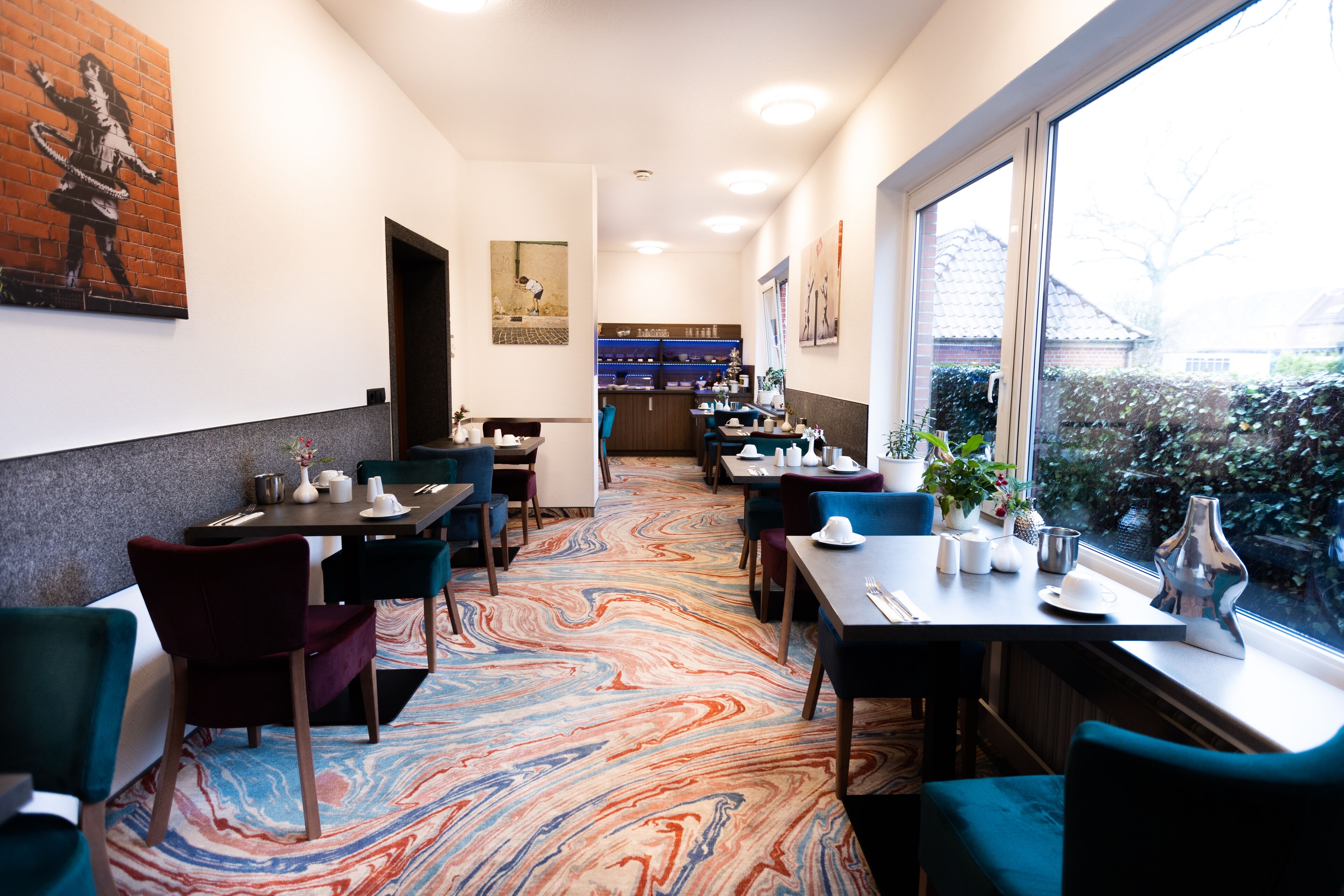 Un'area ristorazione di un hotel con pavimento a motivi marmorizzati, tavoli con sedie e opere d'arte alle pareti.