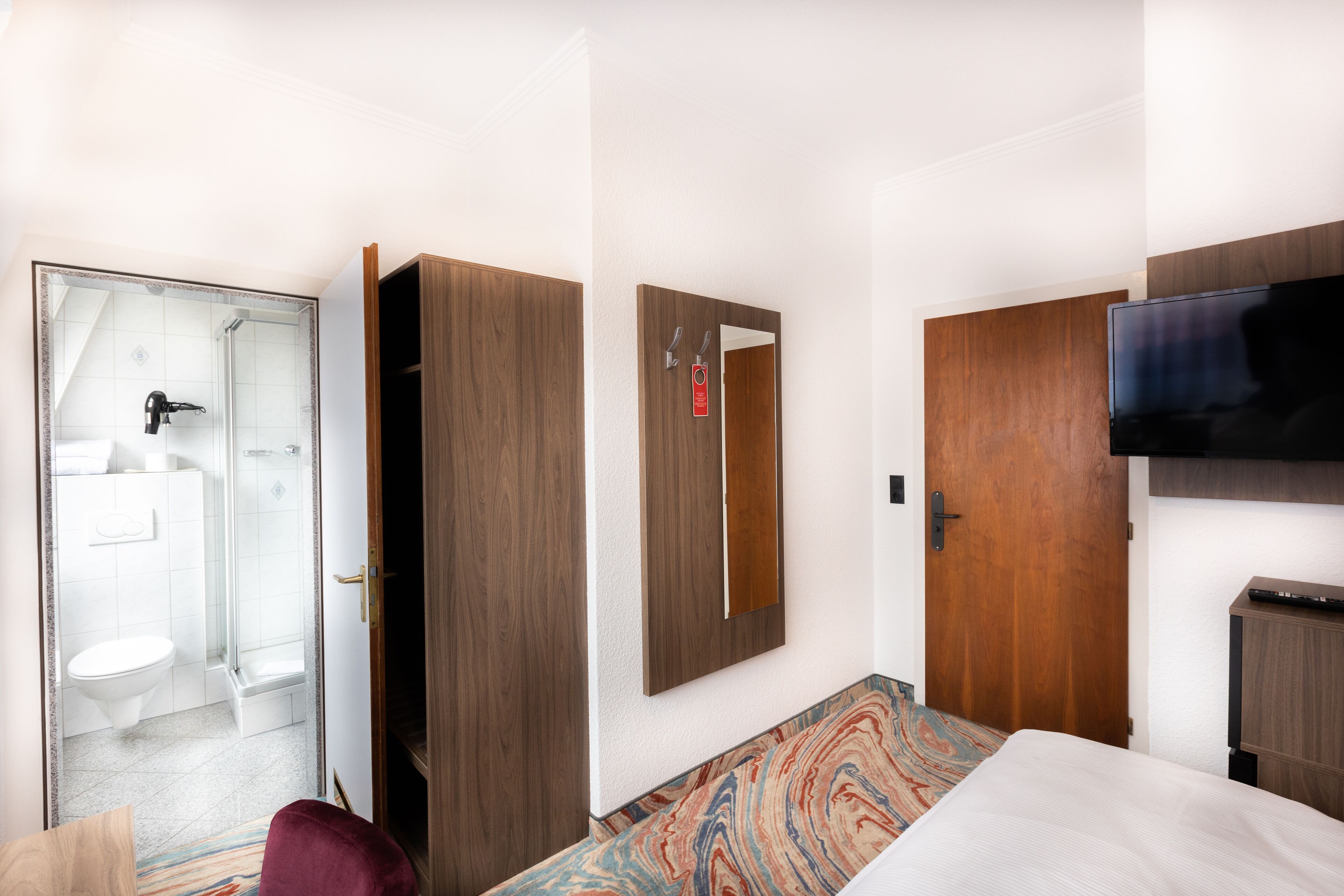 Habitación de hotel con cama, televisión, armario, espejo y baño con ducha e inodoro.