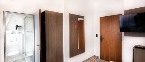 Habitación de hotel con cama, televisión, armario, espejo y baño con ducha e inodoro.
