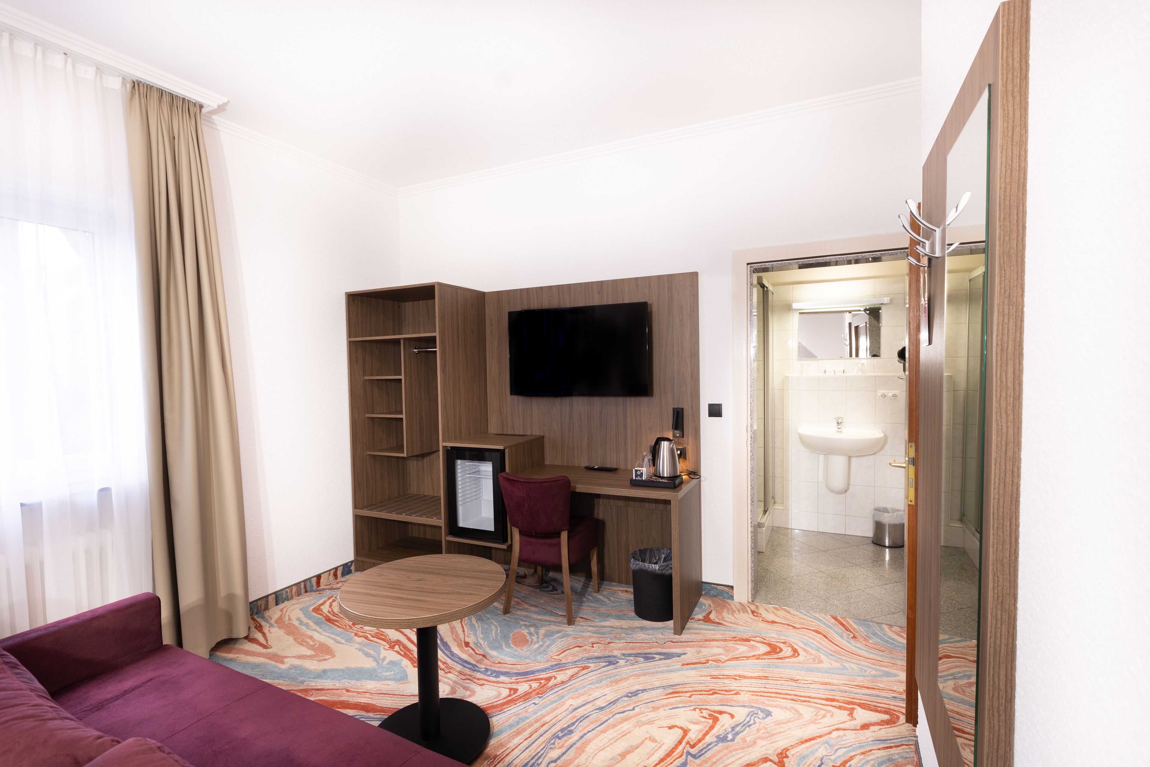 Habitación de hotel con sofá, escritorio con computadora, televisor y baño visible a través de una puerta abierta.