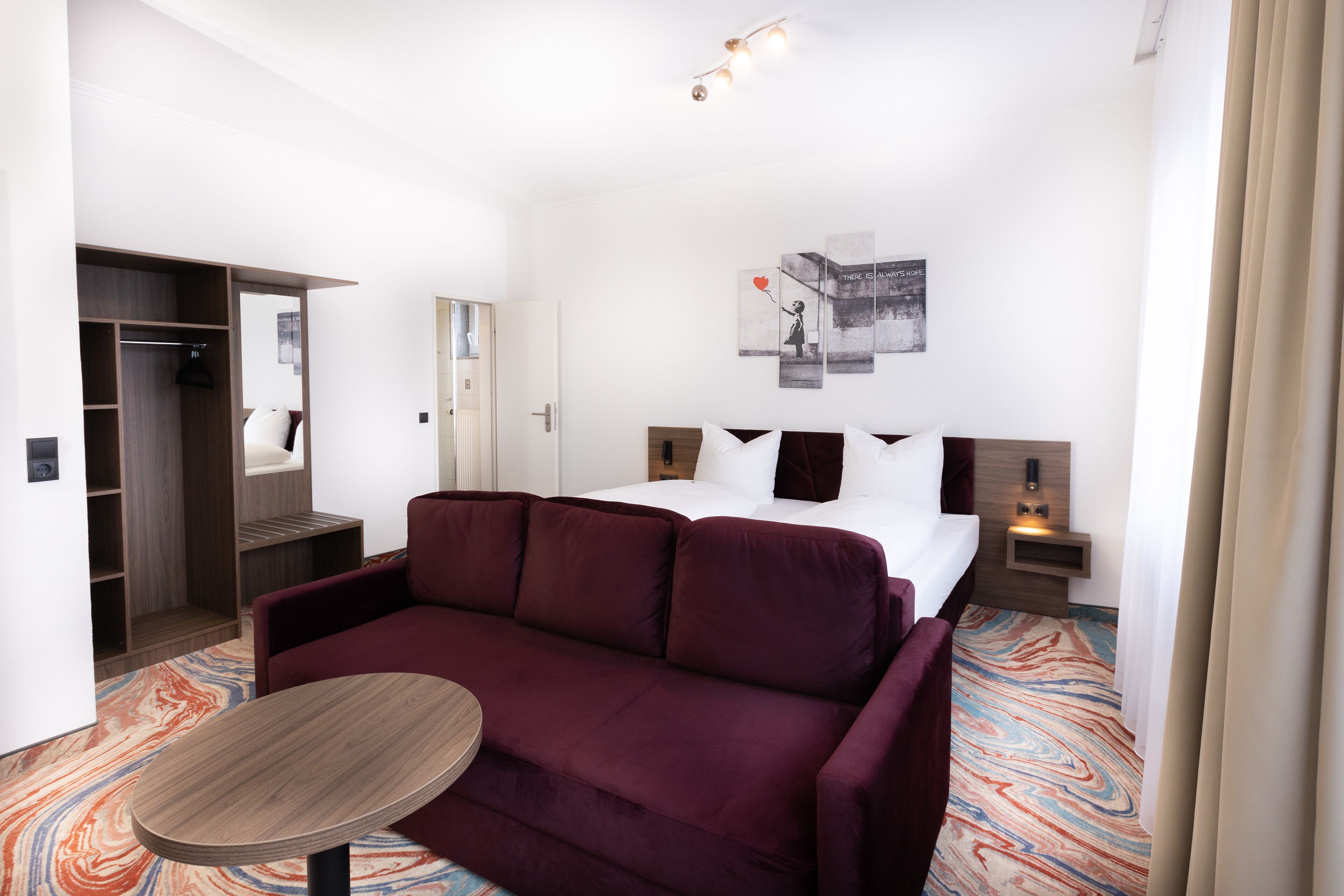 Ein Hotelzimmer mit einem Bett, einem Sofa, einem kleinen Tisch, einem Schrank und einem an der Wand hängenden Kunstwerk.