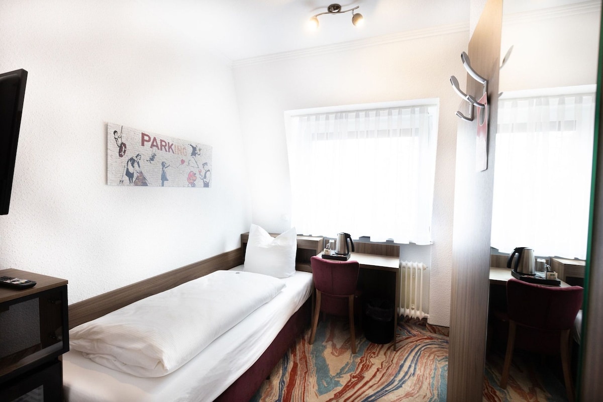 Une chambre d’hôtel avec un lit, un bureau, une chaise, un radiateur et une fenêtre avec des rideaux.