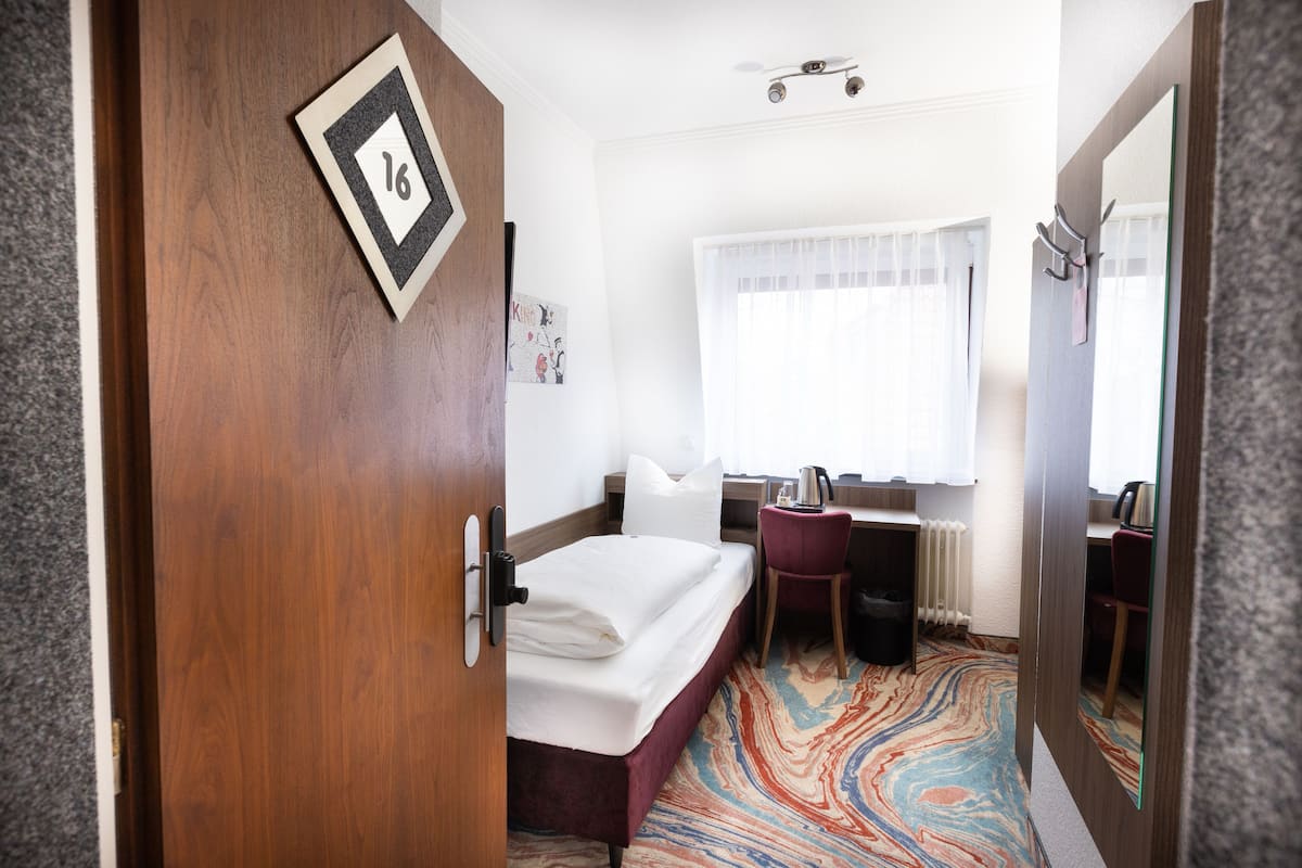 A hotel room with a bed, a desk, a chair, a radiator, and a window with curtains.