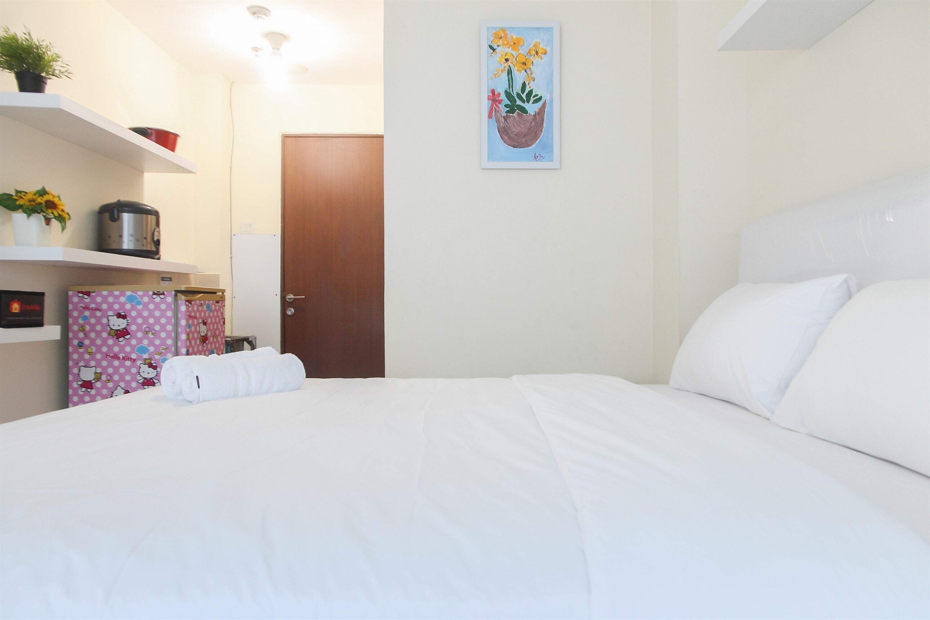 A neatly made bed with white linens, a pink polka-dotted cabinet with Hello Kitty designs, a painting of flowers on the wall, and a wooden door.