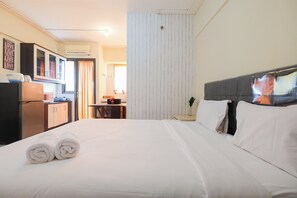 A hotel room with a neatly made bed, a desk with a chair, a wardrobe, and an air conditioning unit on the wall.