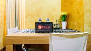 Een hotelkamer met een houten tafel, een zwart-wit tafelonderzetter, een dienblad met travelio snacks en drankjes, een witte vaas met gele bloemen, en een witte stoel.