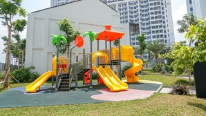 Un parque infantil con toboganes y estructuras para escalar, rodeado de follaje y un edificio al fondo.