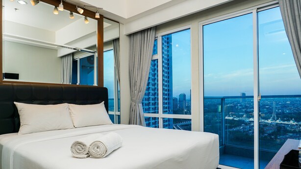 Habitación de hotel con una cama grande, cabecera de madera, mesita de noche y vistas a un paisaje urbano a través de ventanales de piso a techo.