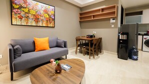 A living room with a gray sofa, a wooden coffee table, a dining table with chairs, a colorful painting, and a kitchen area with a refrigerator and washing machine. - Comfortable and Modern 2BR Menteng Park Apartment (Jakarta)