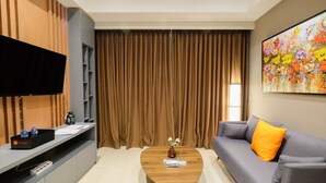 A modern living room with a flat-screen TV, a coffee table with a vase, a sofa with an orange pillow, and a large floral painting on the wall. - Comfortable and Modern 2BR Menteng Park Apartment (Jakarta)