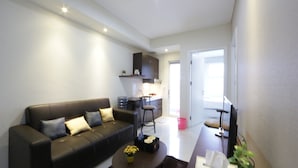 A modern living room with a black leather sofa, a small coffee table with flowers, and a kitchen area in the background. - Clean and Cozy 2BR Apartment @ Parahyangan Residence (Bandung)