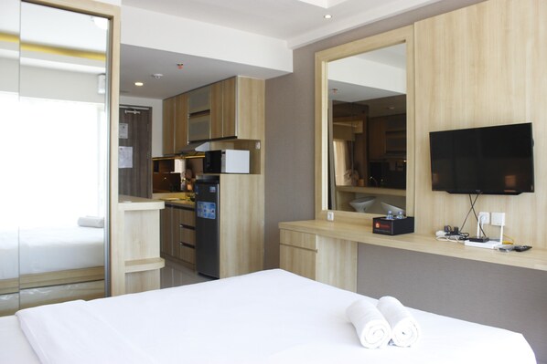 A hotel room with a bed, a large mirror, a TV, a kitchenette, and a bathroom visible through an open door.