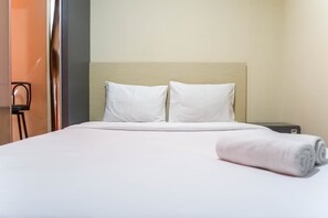 Een net opgemaakt bed met witte lakens en kussens, een opgerolde witte handdoek, en een klein nachtkastje met een lamp.