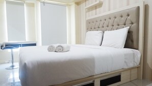Un lit bien fait, avec du linge de lit blanc et deux serviettes enroulées, une tête de lit capitonnée et une table de chevet avec une lampe.