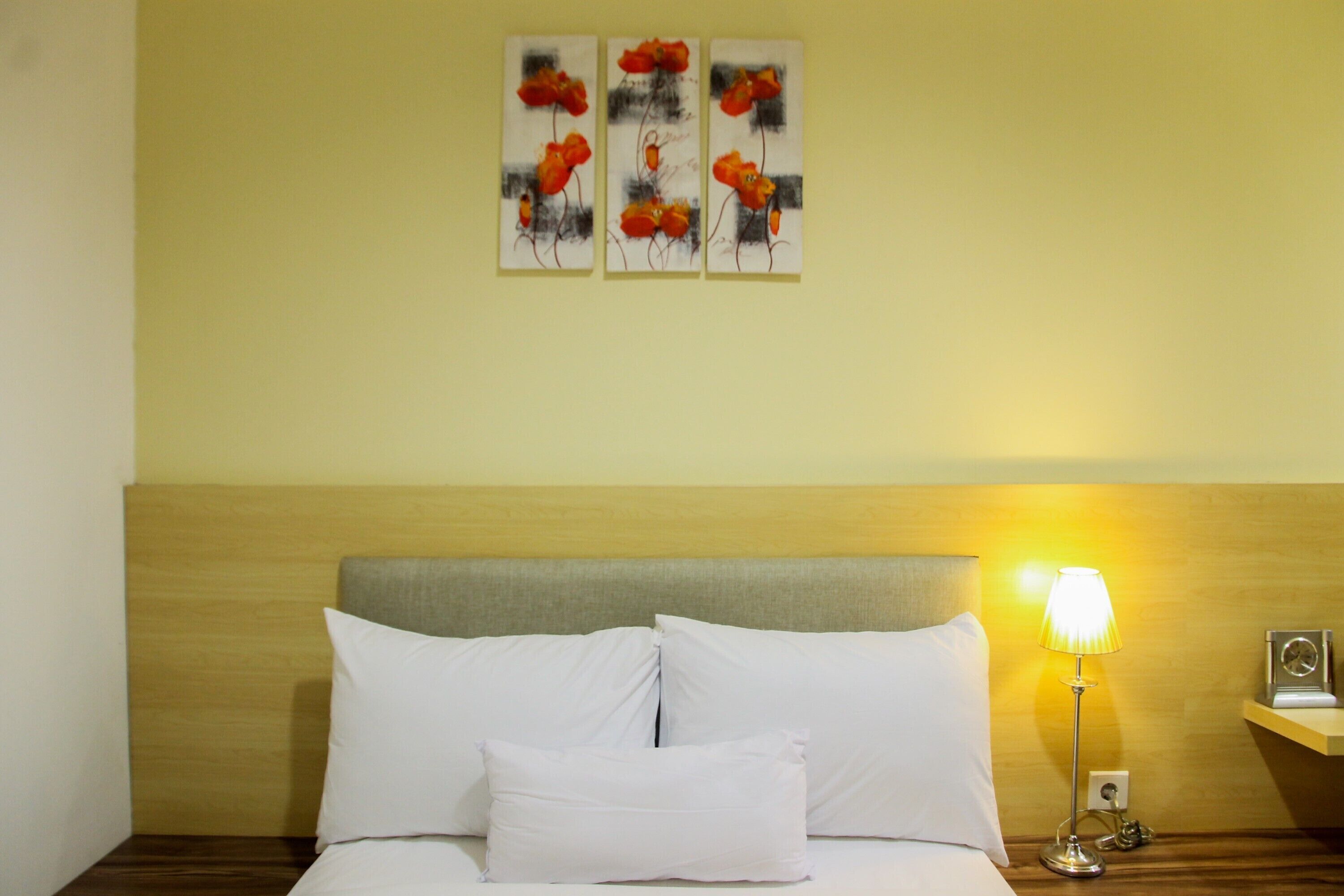 A hotel room with a bed, white pillows, a nightstand with a lamp, and three paintings on the wall.