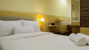 A hotel room with a neatly made bed, a bedside lamp, a small shelf with decorative items, and a mirror on the wall.