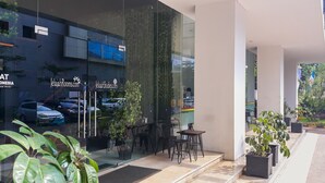 A modern building with glass windows reflecting cars and trees. There are outdoor seating areas with tables and chairs. - Modern Living 1BR @Atlanta Residences Apartment With City View (Depok)