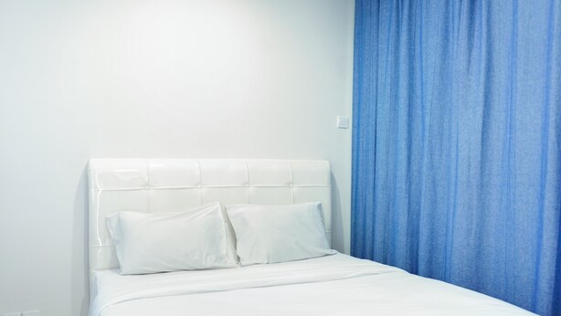 A neatly made bed with white linens and pillows, a blue curtain, and a white headboard. - Modern Living 1BR @Atlanta Residences Apartment With City View (Depok)