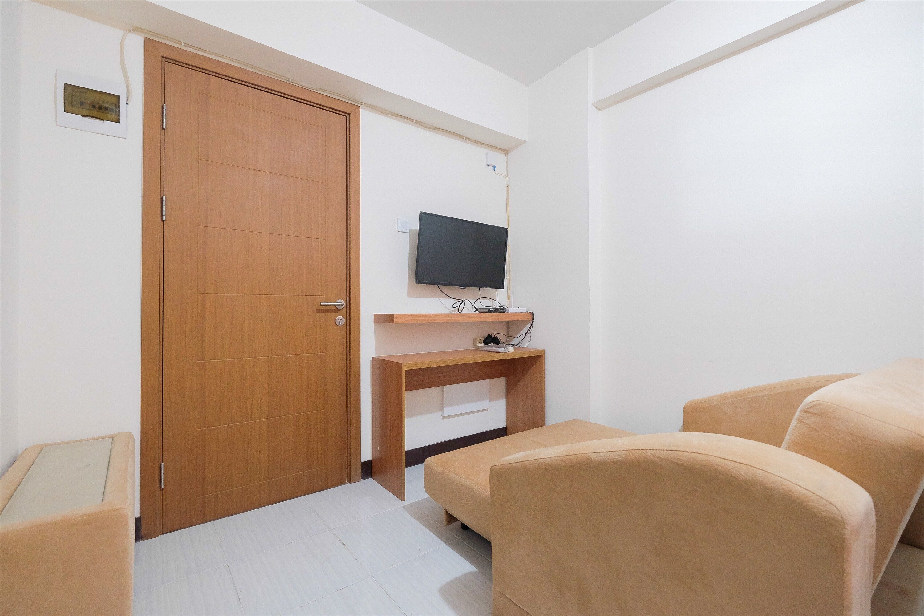 A room with a wooden door, a television mounted on the wall, a wooden desk, and two beige sofas.